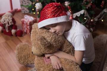  The boy sits near the Christmas tree turned away sad waiting for Santa Claus, close-up