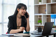 © crizzystudio - Business freelance asian woman talking on phone and working on laptop computer managing her business in workplace, doing business plan on calculator accounting financial report.