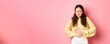 © Mix and Match Studio - Young woman with pain in stomach, holding hands on belly, feeling terrible ache, menstrual cramps, standing against pink background
