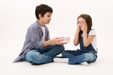 Portrait on white background of adorable teenage boy, loving brother giving a cute present to his younger sister, a little girl covering her eyes. He's greeting her with St Valentine's or women's Day