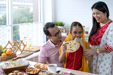 Indian family with their dinner at modern kitchen at home, father mother and daughter having meal together, daughter in traditional dress tasting Indian naan bread, selective focus