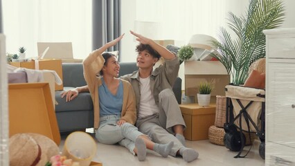 Wall Mural - Happy Asian couple sit on floor in front of sofa couch smile looking at camera making house sign in living room at new house. Husband and wife moving new house, Apartment Rent And Ownership Concept.