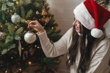 Happy Woman In Cozy Sweater And Santa Hat Decorating Christmas Tree With Stylish Baubles In Atmospheric Festive Room. Merry Christmas! Winter Holidays Preparation