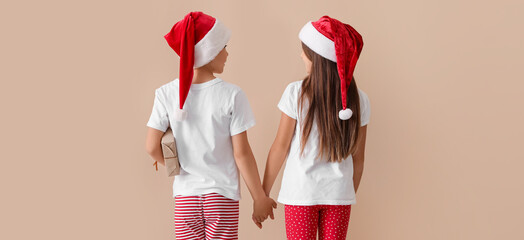 Wall Mural - Little children in pajamas and Santa hats, with Christmas gift holding hands on beige background, back view