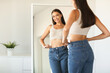 © Prostock-studio - Cheerful young woman wearing jeans and smiling to her reflection in mirror after successful weight loss, copy space