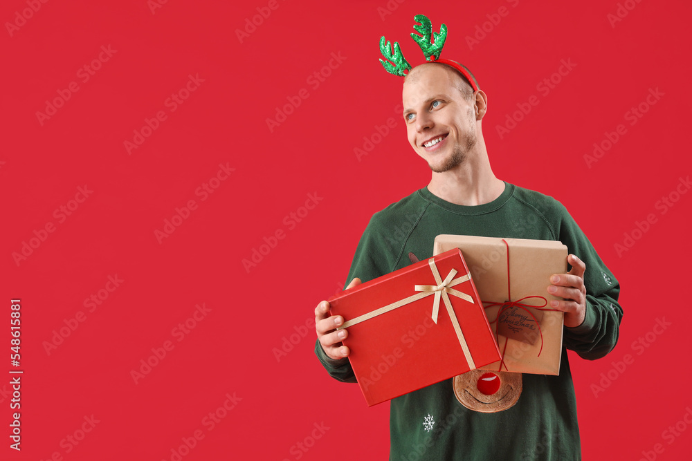 Handsome man in reindeer horns with Christmas presents on red background