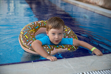 Wall Mural - A little happy boy bathes in a doughnut lifebuoy in clear water in swimming pool in sunny weather