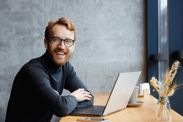 A young red-haired guy, a programmer or an entrepreneur in glasses, in a stylish cafe behind a laptop, looks at the camera and smiles. Freelancer works remotely. Online communication. small business.