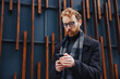 © YURII MASLAK - A young red-haired guy, a businessman in glasses, against the backdrop of urban style, checks mail on a smartphone, communicates in a messenger and drinks coffee. Online communication.