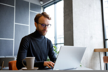 a young red-haired guy, a programmer or entrepreneur in glasses, works in a stylish cafe behind a la