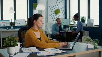 Wall Mural - Focused startup employee typing using laptop keyboard while looking at screen and smiling sitting at desk in busy office. Casual business woman working relaxed with papers with charts and statistics.