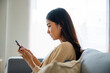 © Monster Ztudio - Happy young asian woman relax on comfortable couch at home texting messaging on smartphone, smiling girl use cellphone chatting, browse wireless internet on gadget, shopping online from home