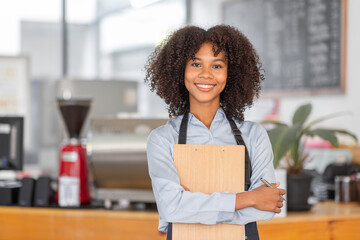 Wall Mural - Female African coffee shop small business owner wearing apron standing in front of counter performing stock check. afro hair employee Barista entrepreneur.