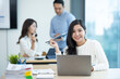 © amnaj - Happy beautiful Asian businesswoman sitting smiling and enjoying working on laptop in office and looking at camera.