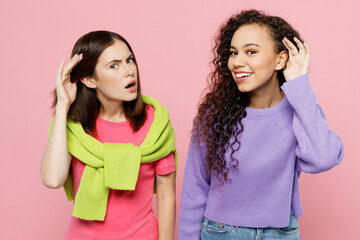 Wall Mural - Young two friends curious nosy women wears green purple shirts looking camera together try to hear you overhear listening intently isolated on pastel plain light pink color background studio portrait.