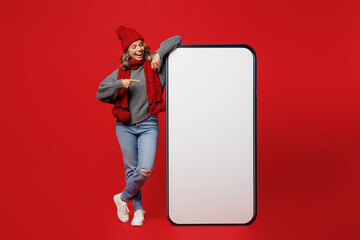 Wall Mural - Full body young woman wear grey sweater scarf hat show big blank screen mobile cell phone with area isolated on plain red background. Healthy lifestyle ill sick disease treatment cold season concept.