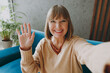 © ViDi Studio - Close up elderly woman 50s year old wear casual clothes sit on blue sofa do selfie shot pov mobile cell phone waving hand stay at home flat rest relax spend free spare time in living room grey wall.