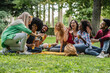 © PintoArt - Multiracial group of friends sitting on the grass in a public park drinking orange juice and waving