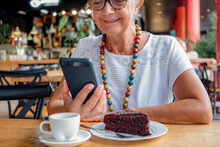 Smiling Senior Woman Relaxing For A Break At Coffee Shop With Chocolate Cake And Coffee Cup. Carefree Elderly Woman With Eyeglasses And Necklace Using Mobile Phone