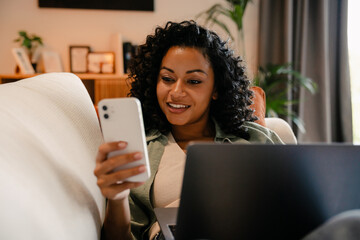 Wall Mural - African woman using smartphone and laptop while lying on sofa in cozy apartment