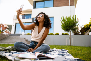 Wall Mural - Young afro woman taking selfie while sitting on grass at backyard outdoors