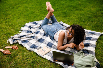 Wall Mural - Young afro woman using laptop while lying on grass outdoors