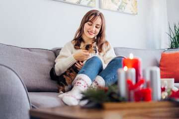 Woman in plaid with tea cup watching movie, TV, petting cat on sofa at home with christmas decoration atmosphere. Lady wear jumper and warm socks. Cozy and comfortable winter concept. Selective focus.