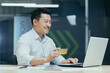 © Liubomir - Online shopping. Satisfied young Asian man sitting in the office at the table with a laptop. He holds a credit card in his hands, makes online purchases, orders through the application, smiles.