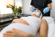 © PoppyPix - young Caucasian pregnant woman lying on a table and a cosmetologist making a facial mask for her, skincare and pregnancy concepts. High quality photo