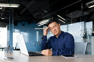 Wall Mural - thinking sad asian man with closed eyes working in office indoors, businessman sitting at desk overworked and frustrated, mature man upset with achievement results.