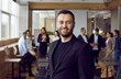 © Studio Romantic - Portrait of smiling business coach after professional corporate training class. Happy satisfied man in suit standing in modern office, with team of company employees in background