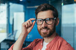 © Liubomir - Closeup photo of young successful architect with beard, mature male designer looking at camera and smiling in glasses and red shirt near window inside office.