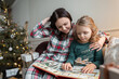 © alones - Happy beautiful mother with daughter sits on sofa and read book near christmas tree with gifts on christmas eve