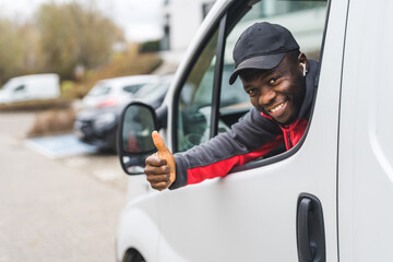 Wall Mural - Black young adult delivery man sitting inside white van leaning out of the windo to look into camera smiling with thumbs up. Horizontal outdoor shot. High quality photo