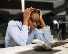 African Young Woman Sitting At Her Desk Clutching Her Head. 