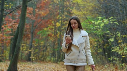 Wall Mural - Walking brunette focused on her phone in autumn park. Long-haired lady taking stroll and looking on the screen of her phone.