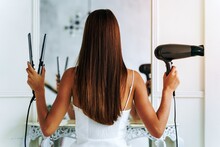 Back View Of The Pretty Young Woman Posing For The Camera With A Hair Dryer And Hair Straightener While Standing In Front To The Mirror