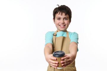 Adorable happy teenage boy wearing blue t-shirt and beige chef's apron, smiling a toothy smile to camera, handing out an eco-friendly cardboard mug of takeaway coffee, isolated over white background