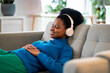 Calm African woman listening to music in wireless headphones lying with closed eyes on sofa. Relaxed black girl in sweater is resting after shopping falling asleep to favorite audio compositions 