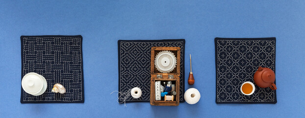 Three handmade napkins embroidered with Sashiko style geometric pattern. Box with sewing accessories and teapot with green tea on blue background. DIY concept. Flat lay, close up, top view, copy space