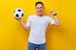 © Bangun Stock Photo - fans excited to support the football sports team.  Asian man 20s wears white t-shirt holding a soccer ball and watching the live stream on tv with doing a winner gesture isolated on yellow background