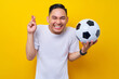 © Bangun Stock Photo - Cheerful young Asian man football fan in a white t-shirt holding a soccer ball and keeping fingers crossed isolated on yellow background. People sport leisure lifestyle concept
