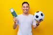 © Bangun Stock Photo - excited young Asian man football fan wearing a white t-shirt holding a soccer ball and passport ticket boarding pass isolated on yellow background. People sport leisure lifestyle concept