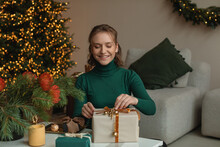 Smiling Young Woman Sitting By A Christmas Tree Wrapping Christmas Gifts