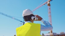 Professional Builder In VR Helmet Standing In Front Of Construction Site And Using Virtual And Augmented Reality Technologies. Office Building And Crane Background. Real Estate And Investment.
