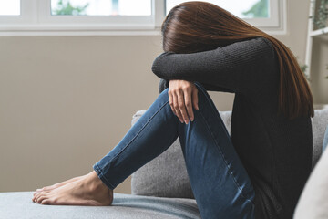 Wall Mural - Unhappy anxiety young Asian woman covering her face with knee on the sofa in the living room at home.