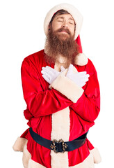 Sticker - Handsome young red head man with long beard wearing santa claus costume smiling with hands on chest with closed eyes and grateful gesture on face. health concept.