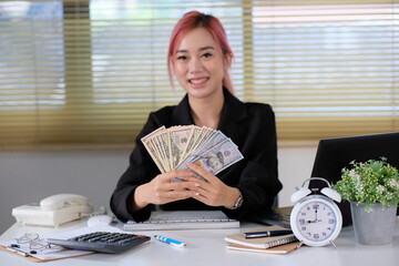 Canvas Print - Businesswoman sits at her desk at work, holding a fan of cash money in dollar banknotes.