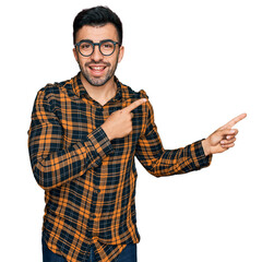 Wall Mural - Hispanic man with beard wearing casual clothes and glasses smiling and looking at the camera pointing with two hands and fingers to the side.