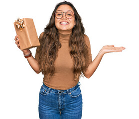 Sticker - Young hispanic girl holding gift celebrating victory with happy smile and winner expression with raised hands